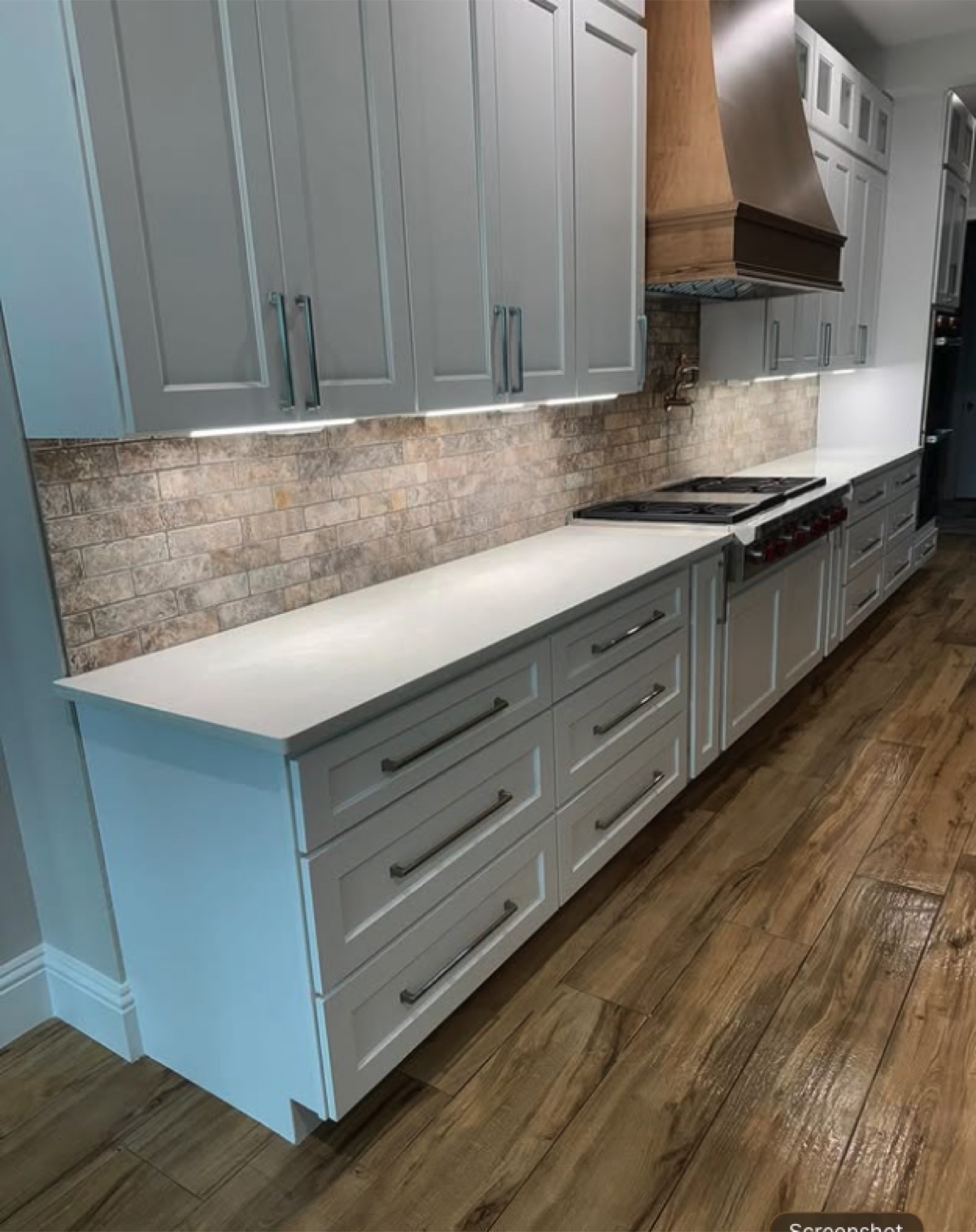 Long white kitchen with stone mosaic backsplash, wood range hood and wide-plank wood flooring