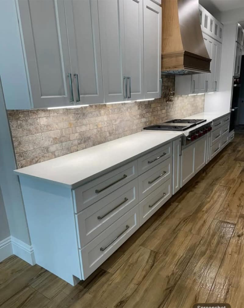 Long white kitchen with stone mosaic backsplash, wood range hood and wide-plank wood flooring