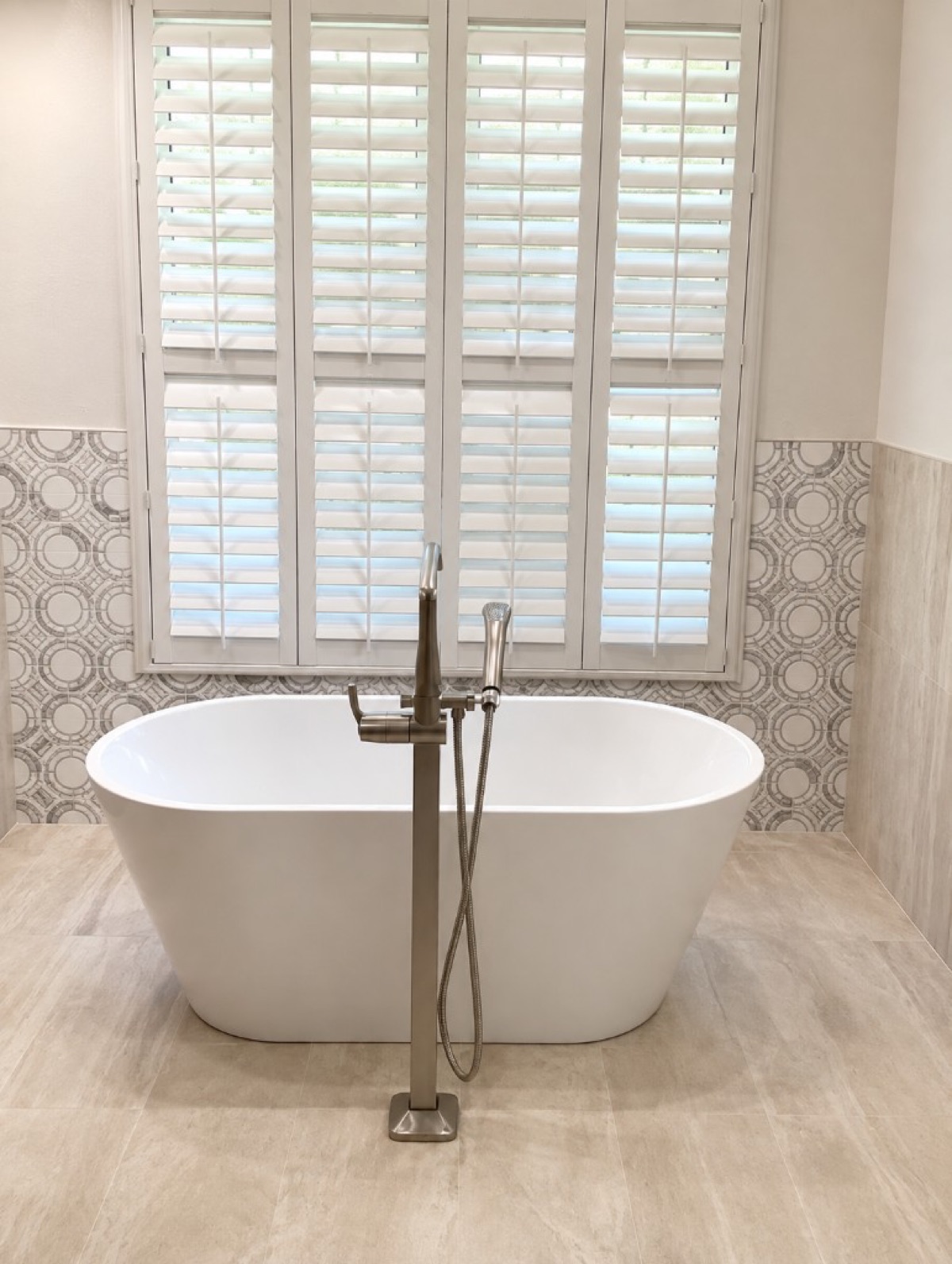 White freestanding soaking tub with a floor-mount filler, circular mosaic wall and plantation shutters