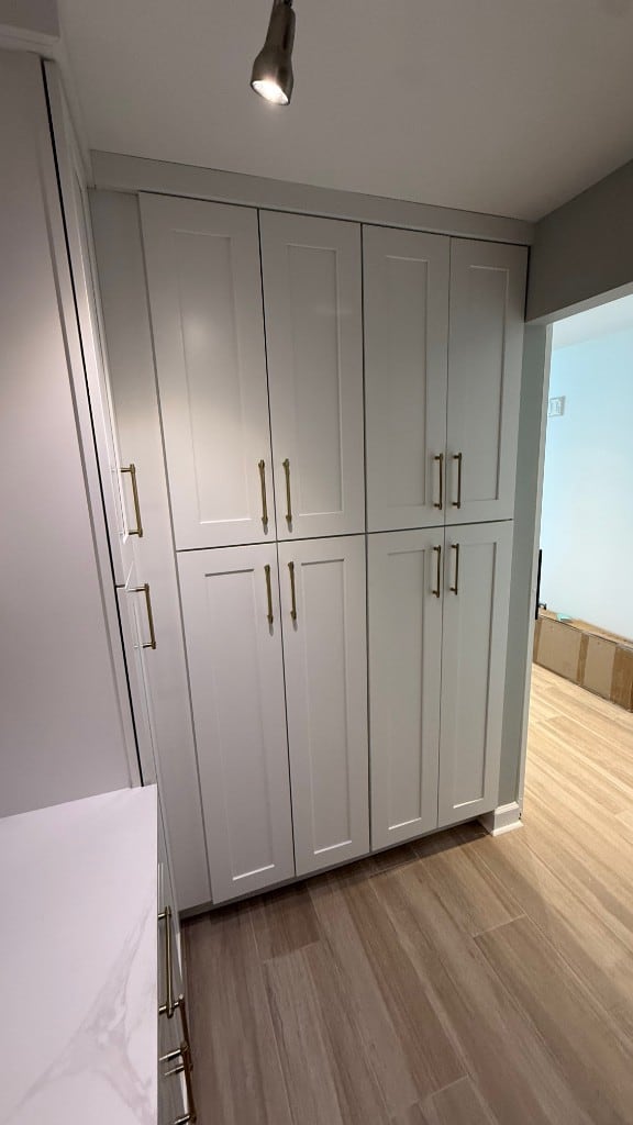Floor-to-ceiling light grey Shaker pantry cabinets with slim gold pulls beside a white quartz countertop and wood-look plank flooring