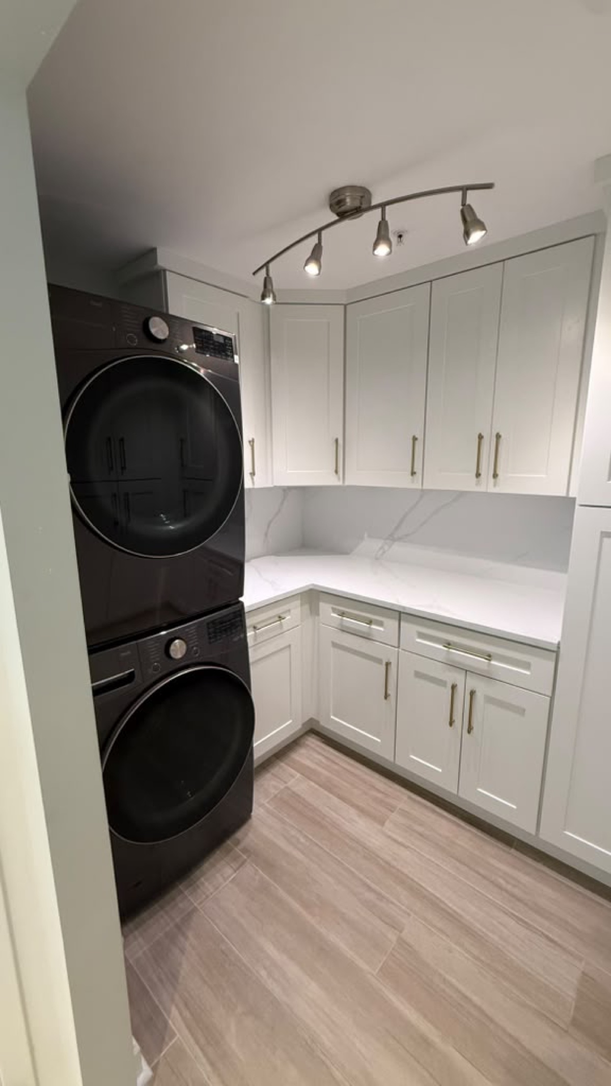 L-shaped laundry with stacked charcoal washer and dryer, white Shaker cabinets, marble-look counters and wood-look plank flooring