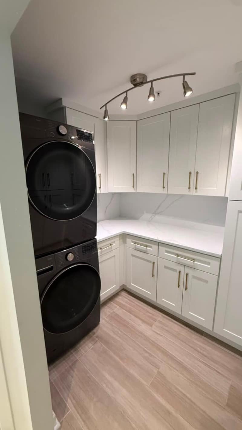 L-shaped laundry with stacked charcoal washer and dryer, white Shaker cabinets, marble-look counters and wood-look plank flooring