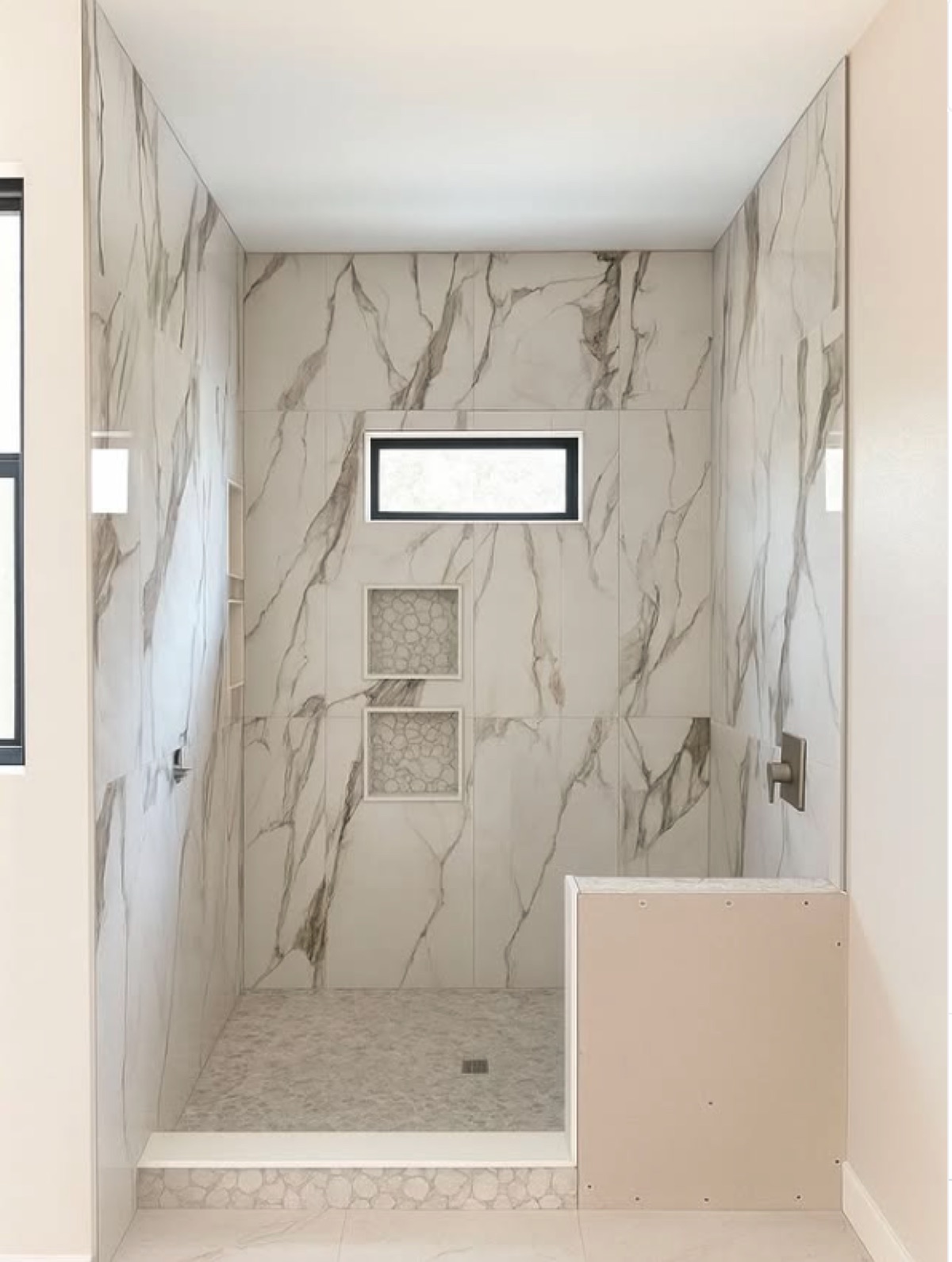 Walk-in shower with marble-look wall tile, pebble mosaic floor and two stacked pebble-lined niches