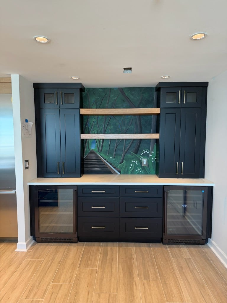 Navy Shaker home bar with gold pulls, white quartz counter, wood floating shelves, a forest mural backsplash and two under-counter beverage coolers