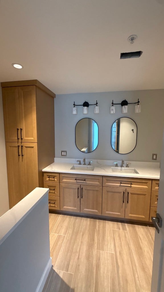 Bathroom double vanity in light oak Shaker cabinets with a white quartz top and black oval mirrors