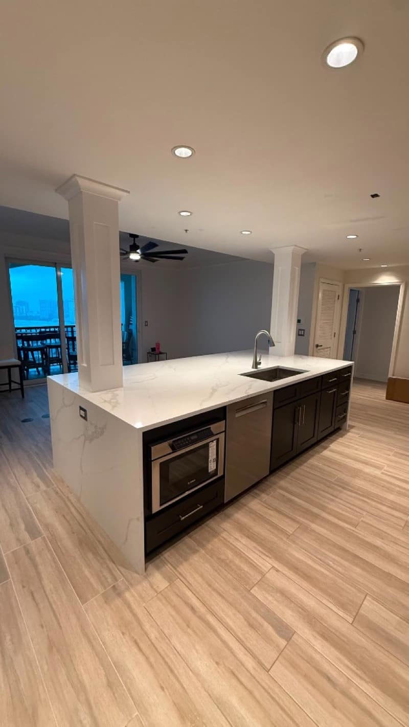 Long kitchen island with a white marble waterfall counter, dark base cabinets, stainless appliances and wood-look floor tile