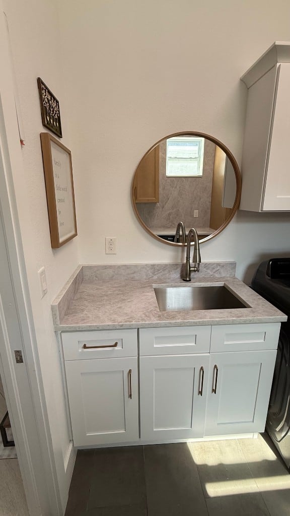 Laundry vanity in white Shaker cabinets with a light grey marble-look counter, stainless undermount sink, brass pulls and a round wood-framed mirror